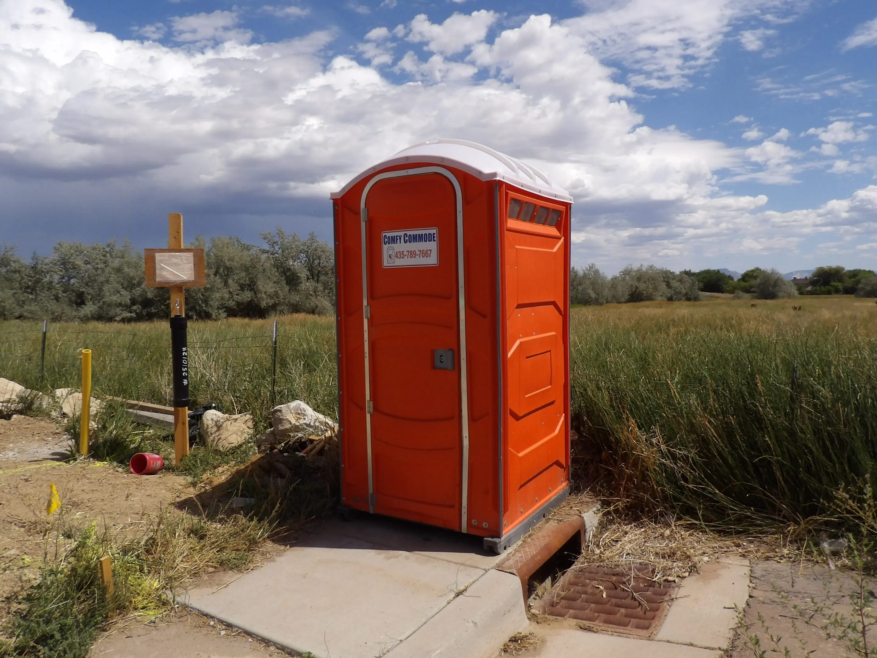 A bright orange portable toilet outdoors with a scenic background.