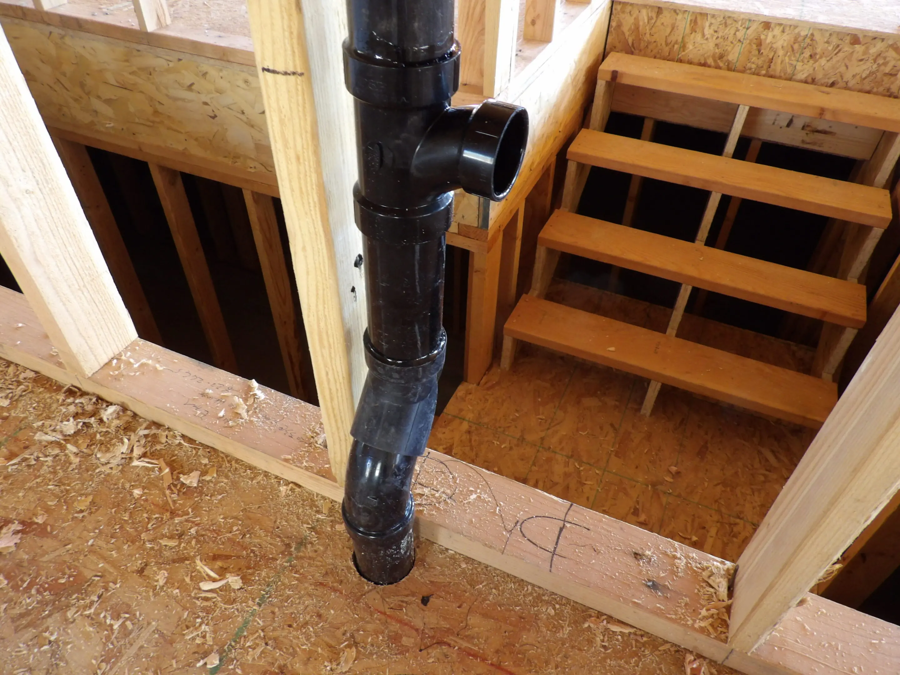 A black PVC pipe installed in wooden floor framing near stairs.